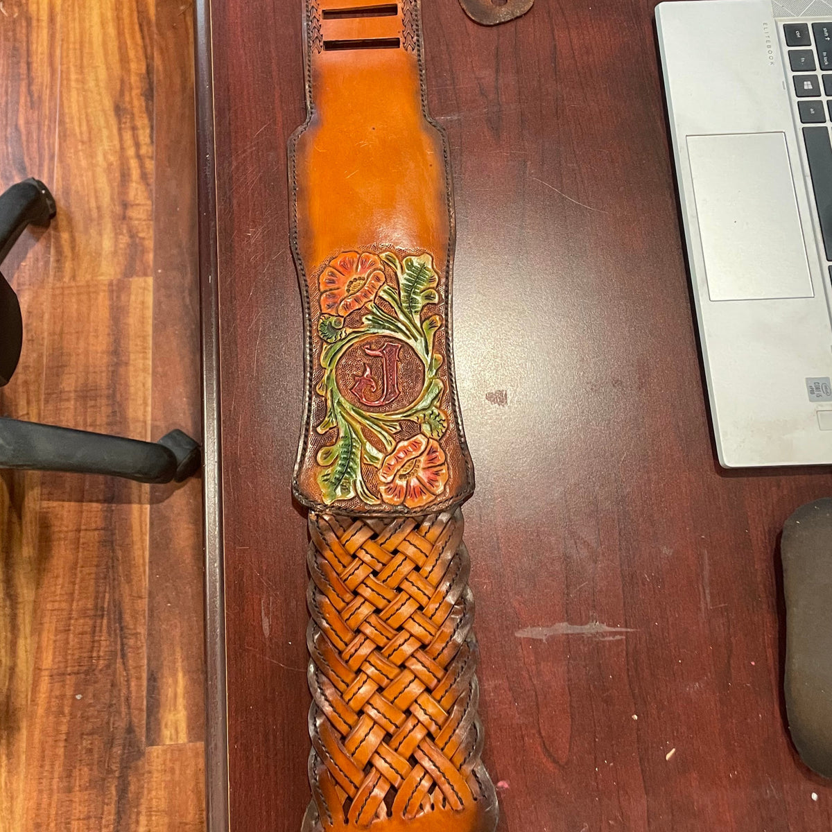 A long, handcrafted leather and woven guitar strap  laid diagonally across a wooden desk, with a laptop, chair, and office floor visible around it.